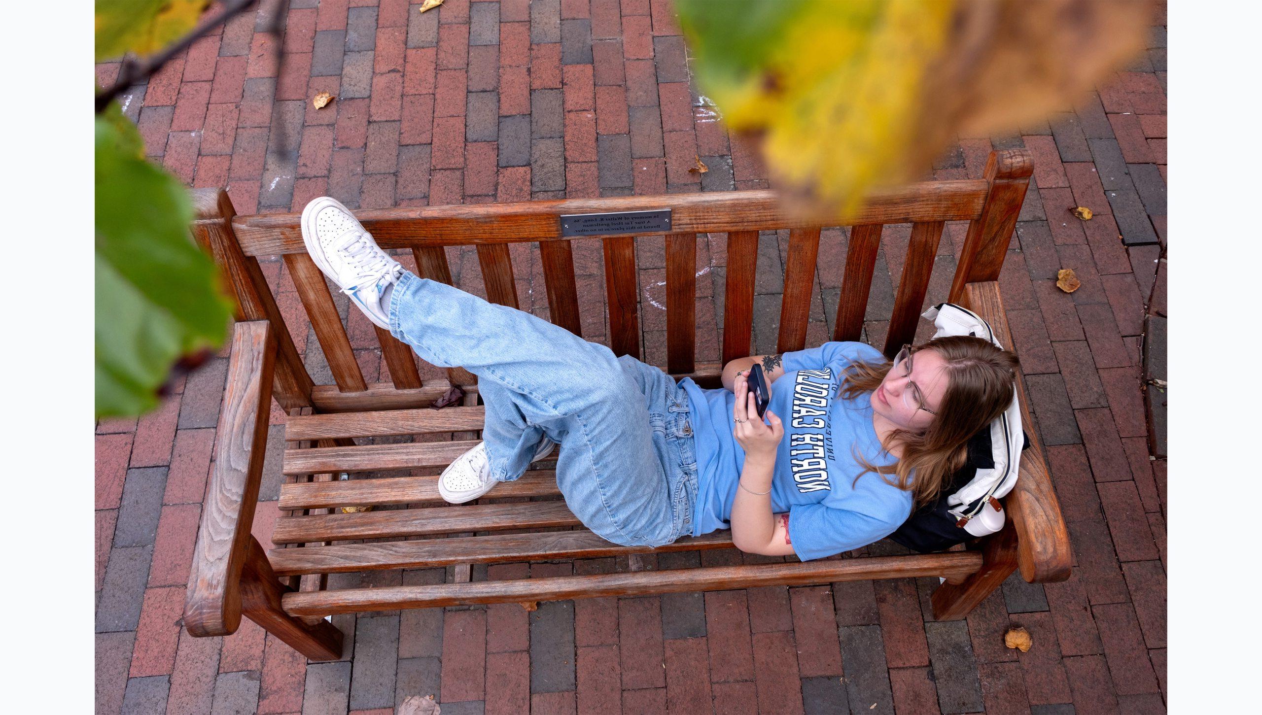 Overhead angle of a student sitting on a wooden bench outside on the campus of UNC-Chapel Hill. Bits of leaves of an overhanging tree are seen as well.